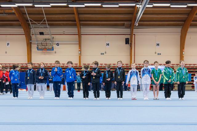 Young gymnasts stand proudly in a row wearing medals at an indoor gymnastics meet, dressed in team uniforms.