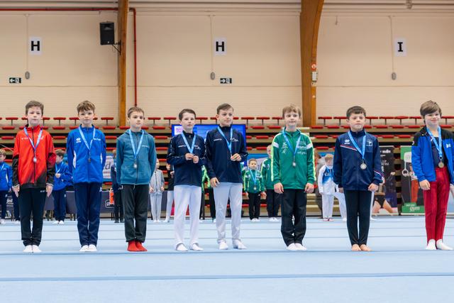 Eight young male gymnasts stand proudly on the competition floor, wearing medals around their necks at an indoor gymnastics event.