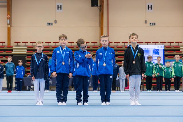 Five young boys wearing bronze medals stand proudly on the competition floor, dressed in blue tracksuits, inside a gymnasium.