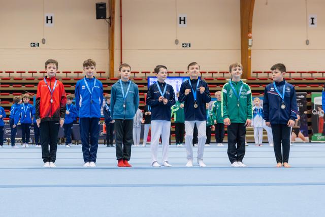Seven young male gymnasts stand proudly on the floor mat displaying their medals at an indoor gymnastics competition.