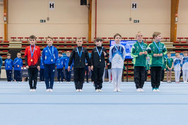 Young boys lined up on the gymnastics floor wearing medals, displaying mixed emotions from proud smiles to neutral expressions.