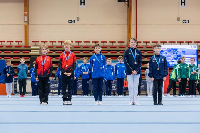 Five young gymnasts stand proudly on the floor podium, wearing medals around their necks at an indoor gymnastics event.