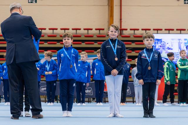 Three young gymnasts stand proudly on the podium wearing bronze medals, while an official presents awards at a gymnastics meet.