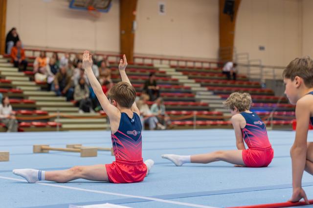 Two boys in matching navy and red leotards perform splits on blue floor mat, one raising arms with pride in a gymnasium.