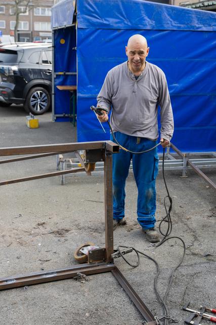 Smiling bald man holds welding equipment beside a metal frame structure at an outdoor worksite with blue tarpaulin.