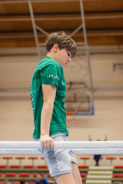Young boy in green t-shirt leans on parallel bar, looking down with focused concentration in a gymnasium.