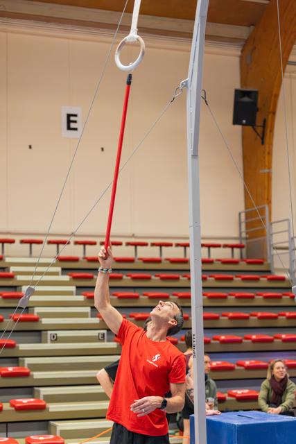 Coach in red shirt uses a long red pole to position gymnastics rings, gazing upward with focus in an indoor arena.