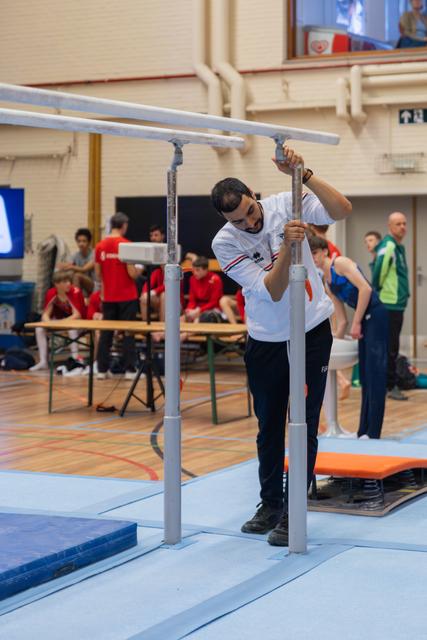 A focused coach adjusts the height of parallel bars in a busy gymnastics hall, athletes in red visible behind.