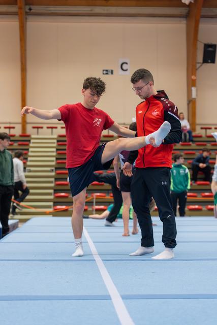 A coach in red steadies a young gymnast practicing a leg raise on the blue floor mat in a gymnastics hall.