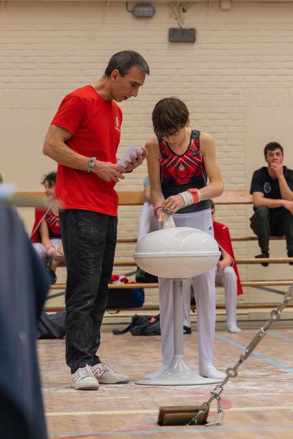 A focused coach in red helps a young gymnast apply chalk to his wrists before a pommel horse routine in a gymnasium.