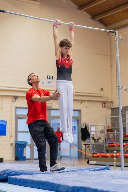 A coach steadies a young male gymnast hanging from the high bar, looking up attentively in an indoor gymnastics hall.
