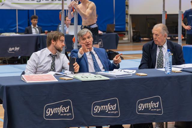 Three judges in suits discuss intently at a Gymfed table, center judge gesturing expressively while holding a phone.