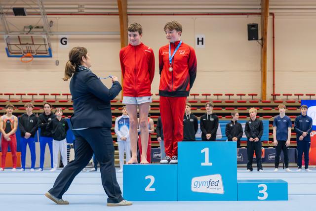 Woman pins a medal on a smiling boy in red on the 1st place podium, as his teammate stands on 2nd.