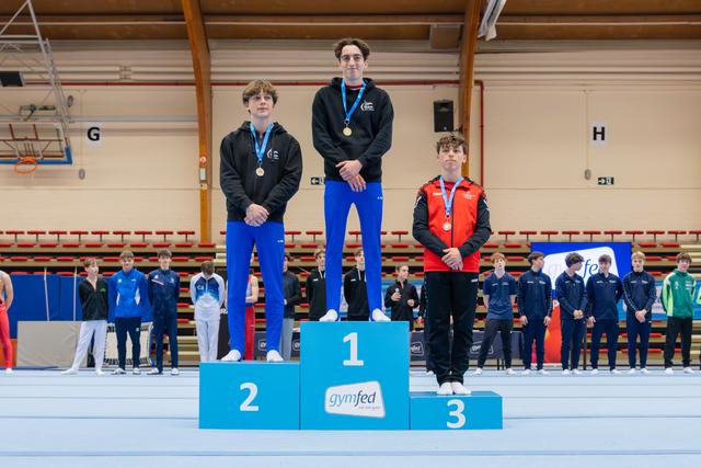 Three young gymnasts stand on a Gymfed podium; the gold medalist stands tall in the center, flanked by silver and bronze winners.