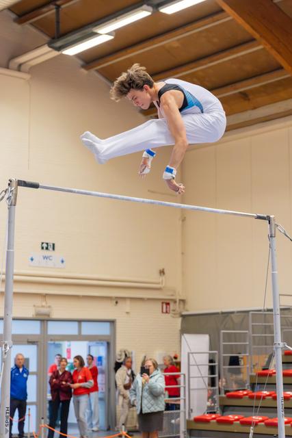 Male gymnast soars above the high bar in a tucked position, body coiled with focus and control during indoor competition.
