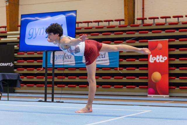 Young male gymnast holds a precise arabesque balance on the floor, arms extended, in a competitive gymnastics hall.