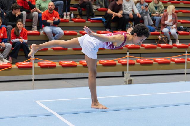 Young gymnast holds a precise arabesque on the floor, arms extended, focused expression, spectators watching in bleachers behind.