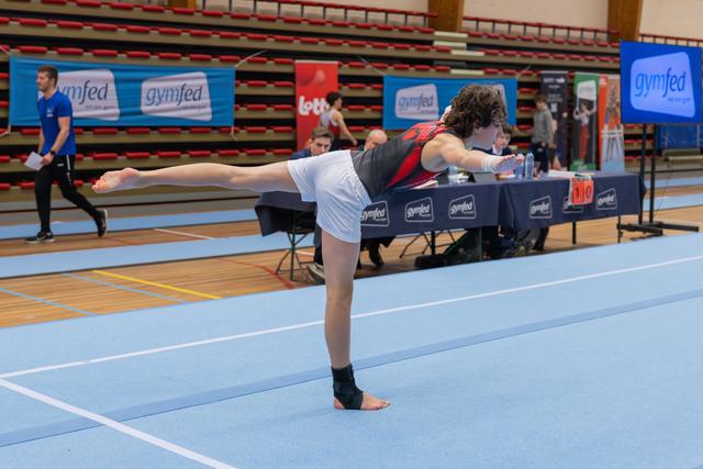 Young gymnast holds a precise arabesque on one leg during a Gymfed floor routine, arms extended forward with focus.