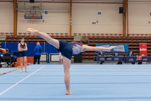 Gymnast performs a precise arabesque on the floor, arms extended, leg raised high, in a competitive gymnastics hall.