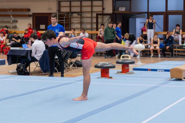 Young male gymnast holds a precise arabesque balance on the blue floor mat during an indoor gymnastics competition.