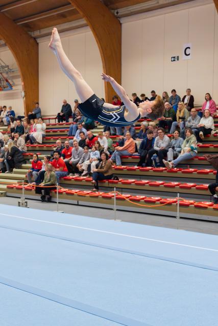 Young gymnast in navy leotard executes a soaring back layout, legs split, arching gracefully above the blue competition floor.
