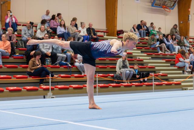 Young male gymnast holds a precise arabesque balance on the floor, focused expression, during a gymnastics meet with spectators.