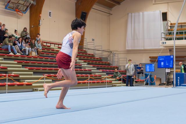 Young male gymnast sprints barefoot across the blue floor mat, focused and poised, in a packed indoor gymnastics hall.