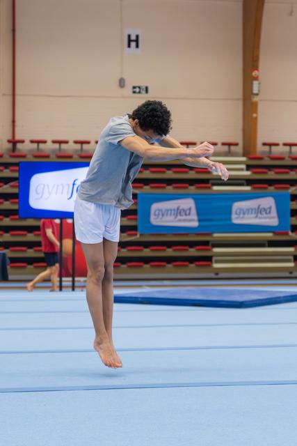 Young male gymnast leaps mid-air with arms tucked forward, focused expression, on blue floor at Gymfed competition.