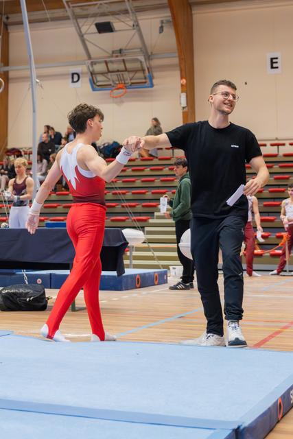 Young male gymnast in red leotard shakes hands with smiling coach on floor mat in a gymnastics hall.