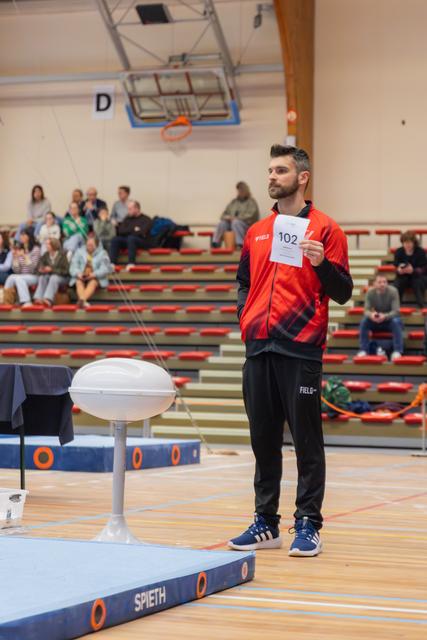 Bearded man in red FIELD jacket holds number 102 card, standing confidently beside a vault in a gymnasium with spectators.