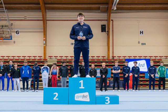 Young male gymnast stands proudly on the first-place podium, holding a trophy at a Gymfed gymnastics competition.