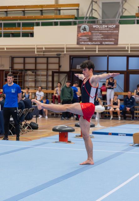 Young male gymnast holds a standing scale pose on blue floor mat, arms outstretched, expression focused and controlled.