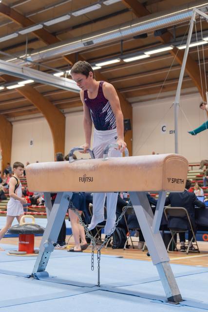 Young male gymnast grips pommel horse handles, concentrating intensely before his routine in a busy gymnastics hall.