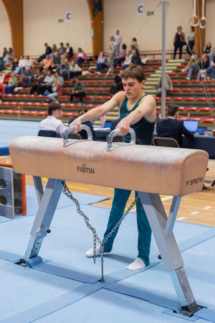 Young male gymnast grips pommel horse handles with focused concentration, preparing his routine at a busy gymnastics meet.