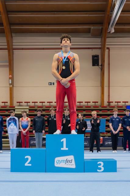 Young male gymnast stands tall on the 1st place podium, chin raised, gold medal around his neck at a Gymfed event.