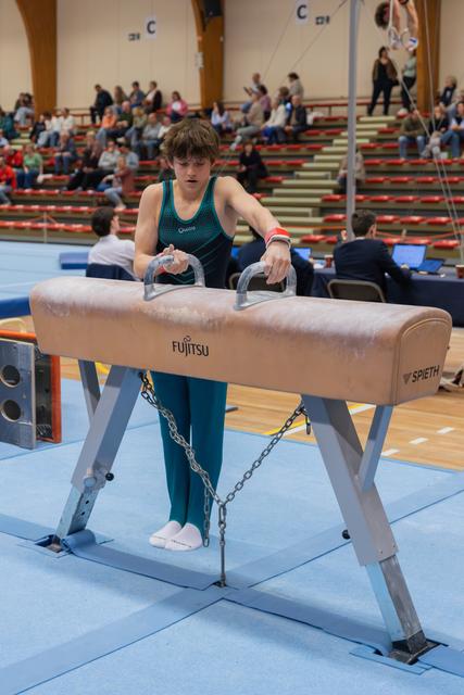 Young male gymnast stands focused at a Fujitsu-branded pommel horse, gripping the handles before his routine in a packed gym.