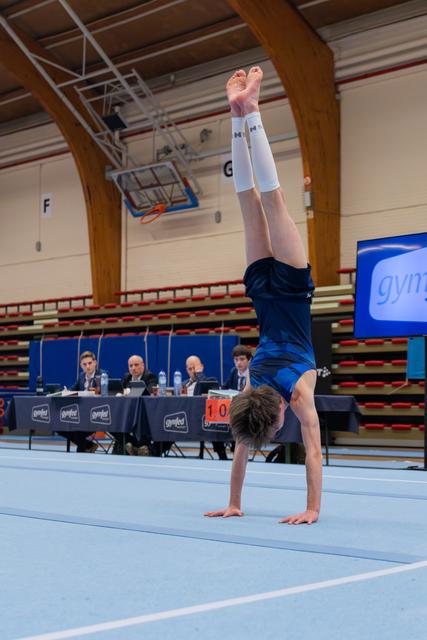 Young male gymnast holds a perfect handstand on floor, legs split wide, judges watching from table behind.