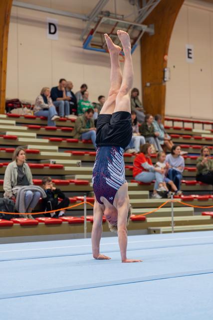 Young gymnast performs a perfect handstand on the floor mat, legs straight up, competing before a seated crowd.