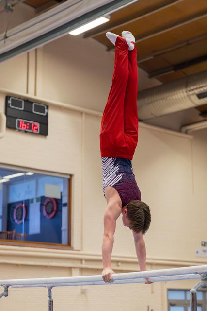 Young male gymnast performs a precise handstand on parallel bars, legs straight in red leotard, in an indoor gym.