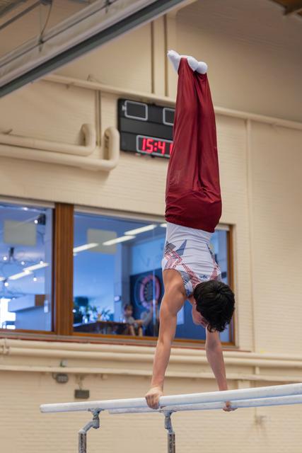 Young male gymnast holds a perfect handstand on parallel bars, legs vertical in red trousers, during an indoor competition.