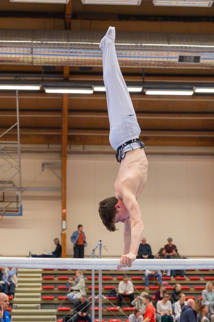 Shirtless male gymnast performs a perfect handstand on parallel bars, legs split upward, in a packed indoor gymnasium.