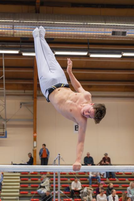 Young male gymnast holds an impressive handstand on parallel bars, body fully extended, in a packed indoor gymnasium.