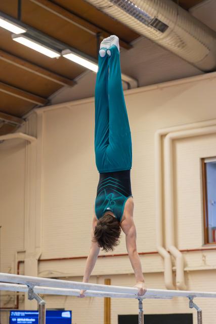Young gymnast performs a perfect handstand on parallel bars, body fully extended upward in teal leotard and warmup top.