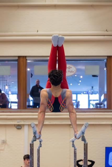 Male gymnast holds a perfect handstand on parallel bars, legs straight up, wearing red leotard in indoor gym.