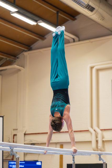 Young male gymnast holds a perfect handstand on parallel bars, body fully extended, wearing a teal leotard in an indoor gym.
