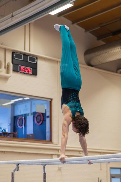 Young male gymnast holds a perfect handstand on parallel bars, legs straight up, wearing a teal leotard in an indoor gym.