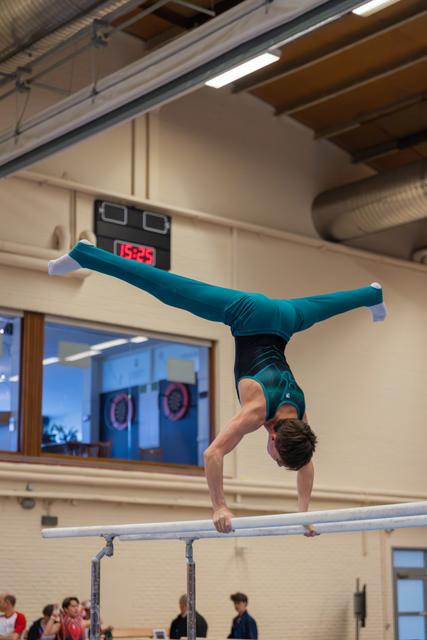 Male gymnast performs a handstand with legs split on parallel bars, wearing a teal leotard, in an indoor gymnasium.