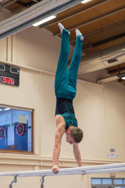 Young male gymnast holds a perfect handstand on parallel bars, legs split, in a gym with a digital clock visible.