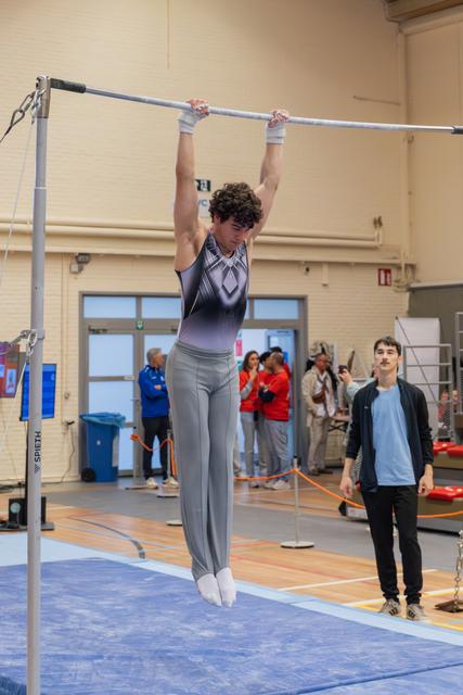 Young male gymnast hangs from a high bar, body fully extended, focused expression, while a teammate watches nearby.