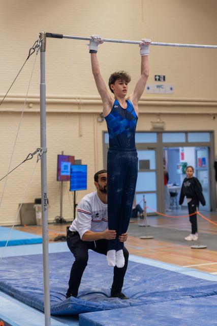 Young male gymnast hangs from a high bar while his coach steadies his legs, focused and composed in an indoor gym.